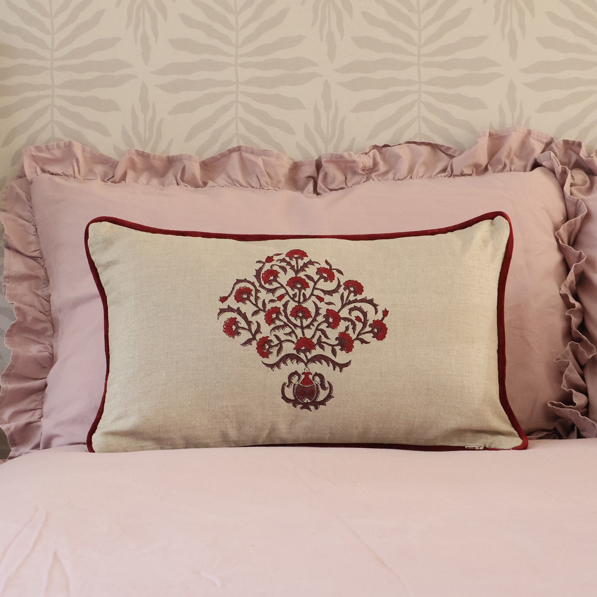 Mulberry Mudita Cushion in natural linen with deep red embroidered floral motif and red velvet piping, styled on a pink ruffled pillow.